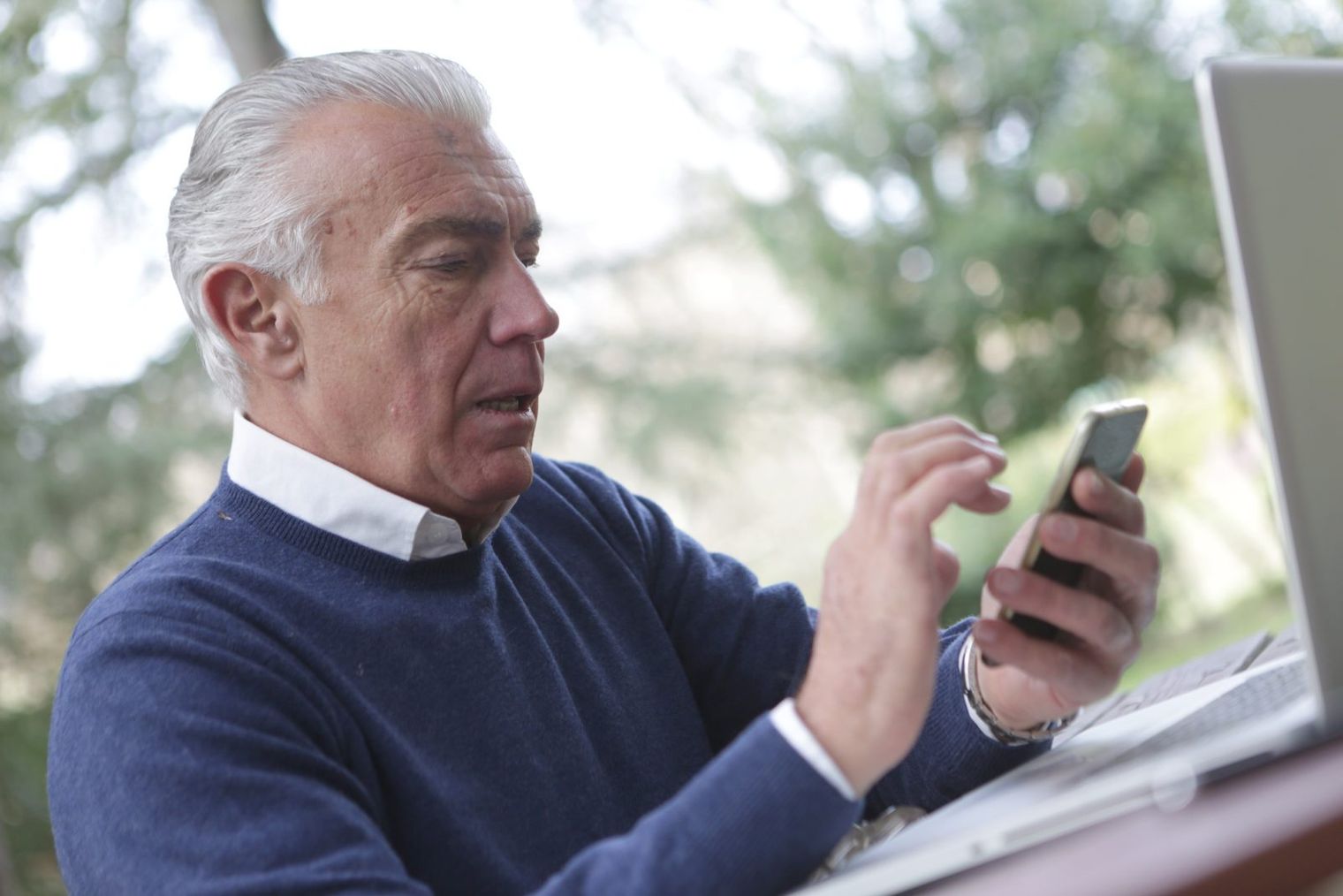 A man with gray hair in a blue sweater using a smartphone at an outdoor table, with a laptop beside him.