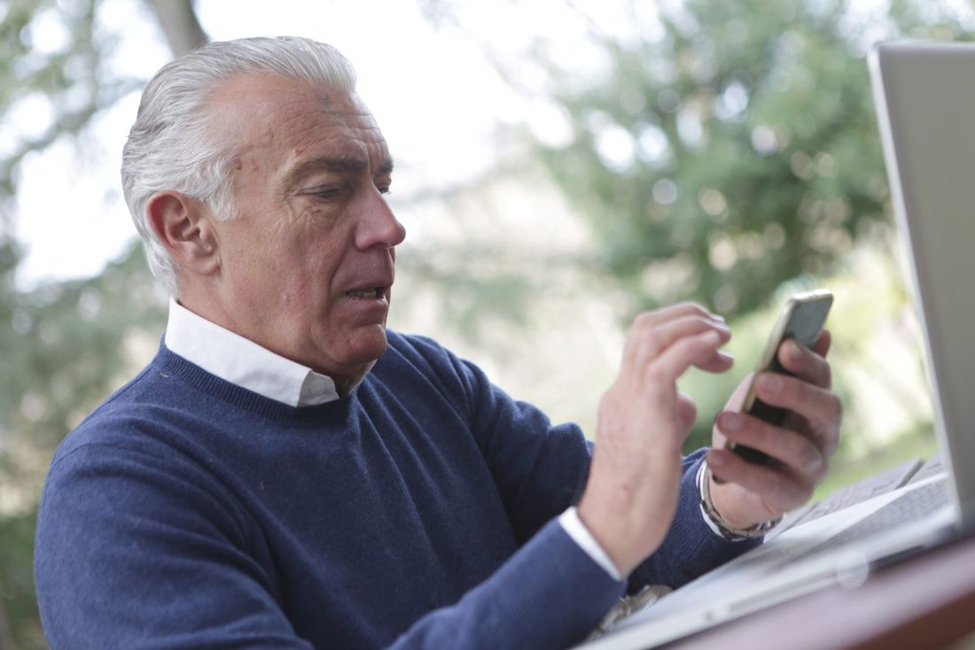 A man with gray hair in a blue sweater using a smartphone at an outdoor table, with a laptop beside him.